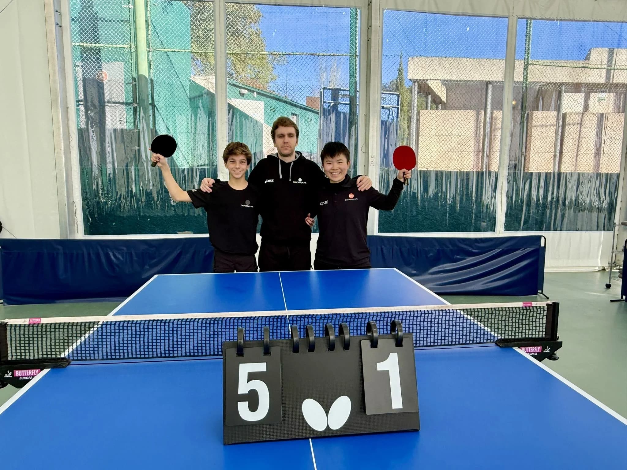 Tres jugadores del equipo de competición de tenis de mesa del Club Deportivo Bonasport levantan sus palas detrás de la mesa de juego